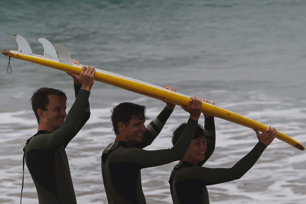 3 Surfer tragen ein Surfbrett und lachen dabei