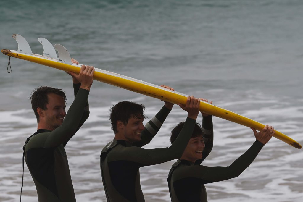 3 surfers carrying a surfboard over their heads laughing